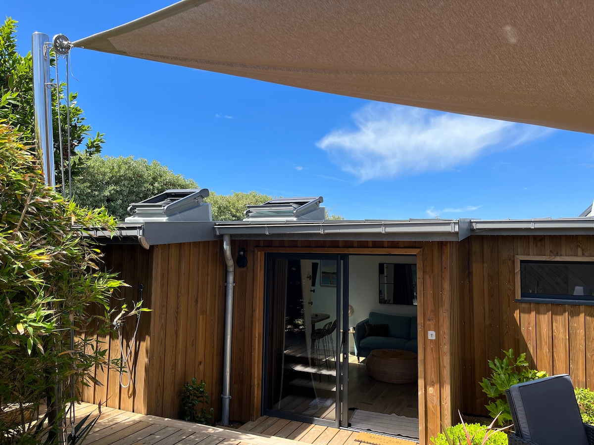 An outdoor terrace is shaded by a large canopy, providing protection from the sun. Wooden decking leads to a modern entrance with glass doors, revealing glimpses of the living area inside. Surrounding greenery adds a touch of nature to the setting.