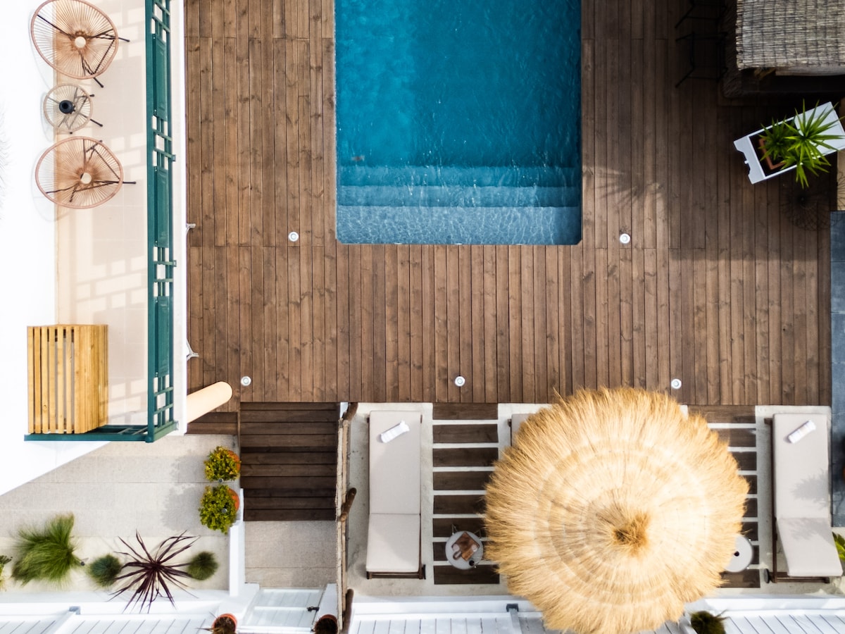An aerial view captures the inviting pool surrounded by wooden decking and lounge areas. Sunbeds are positioned strategically, and a large, round straw umbrella offers shade. Potted plants add greenery to the outdoor space, creating a relaxing atmosphere ideal for guests.