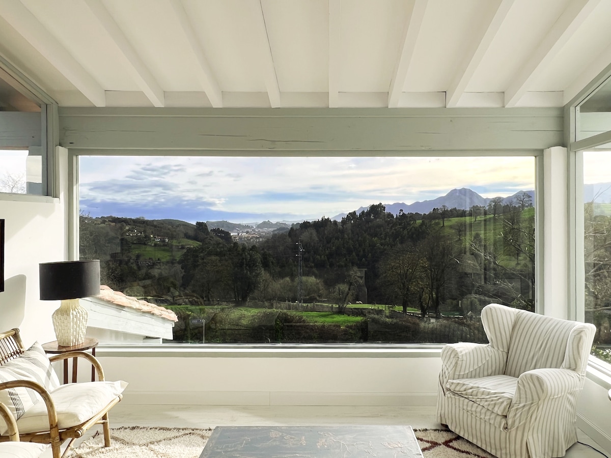 A spacious living area is highlighted by large windows that provide sweeping views of the surrounding landscape, featuring green hills and distant mountains. Natural light fills the room, which includes a comfortable armchair and a striped sofa, fostering a relaxed atmosphere.