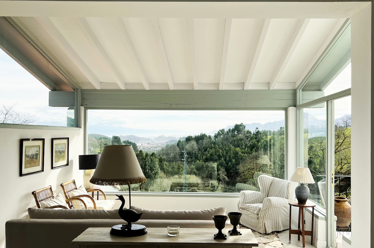 A bright living area features large glass windows offering expansive views of the green landscape. A light sofa is complemented by a striped armchair. A wooden table holds a lamp, vases, and decor, enhancing the serene ambiance of the space.