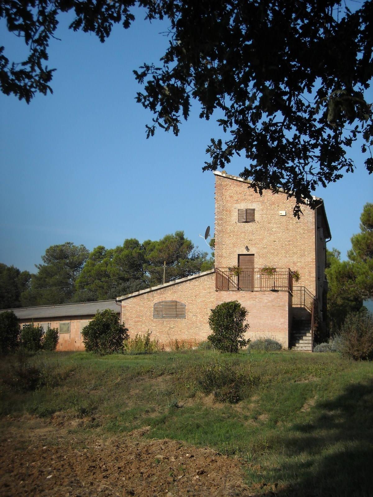 A two-story brick building is set amidst lush greenery, with access stairs leading to a balcony. The exterior features several windows and satellite dish, while surrounding fields provide a peaceful rural landscape.