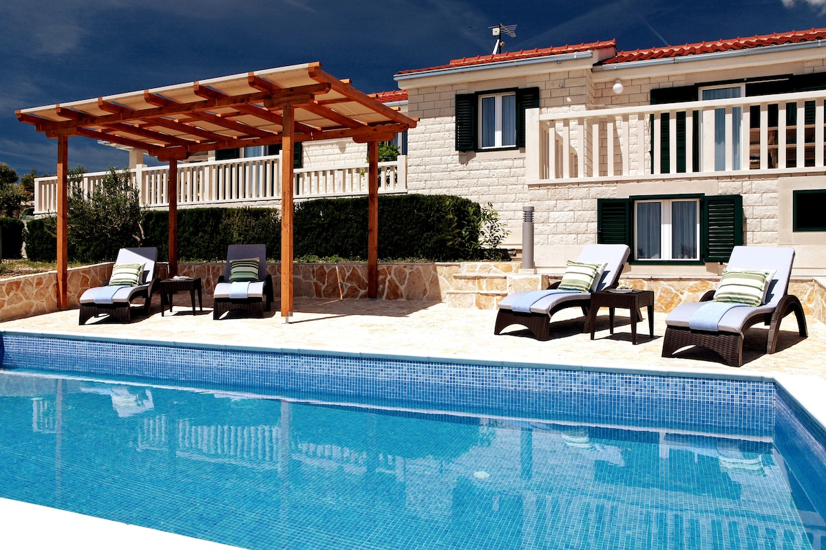 A private pool surrounded by stone tiles reflects serene blue water. Two loungers with light cushions and green accents are positioned under a wooden pergola. The villa's exterior exhibits a pale stone façade and white railings, creating a welcoming outdoor space.