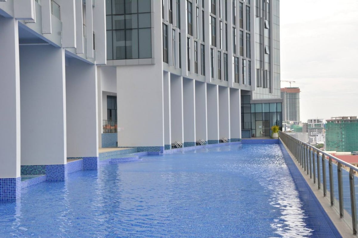 An expansive outdoor swimming pool is visible, featuring a serene blue surface reflecting the sky. The pool area is surrounded by tall, modern buildings, with a railing providing safety along the pool's edge.