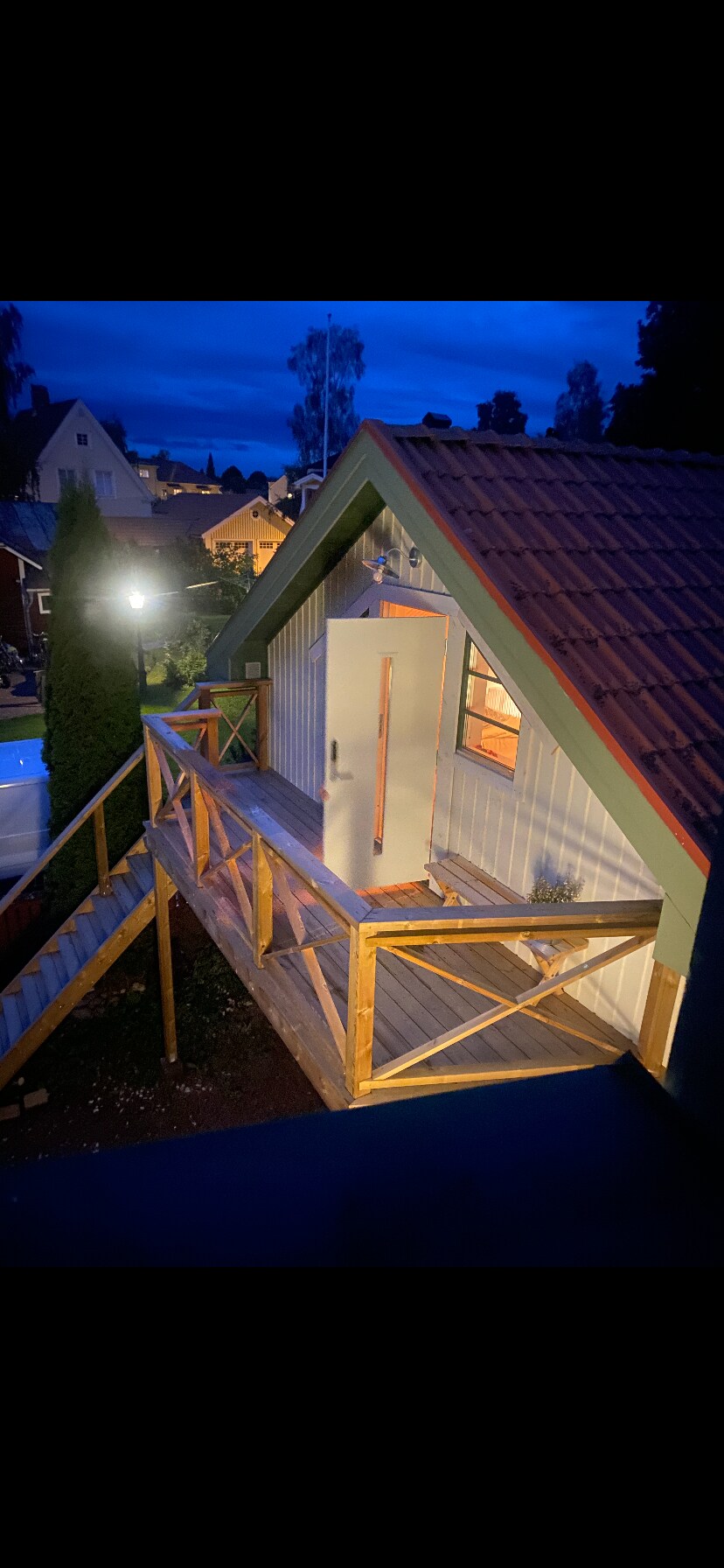 An exterior view of the loft is showcased during twilight, featuring a wooden staircase leading to a bright entry door. The surrounding area is illuminated by soft outdoor lighting, complementing the cozy structure amid nearby homes and greenery.