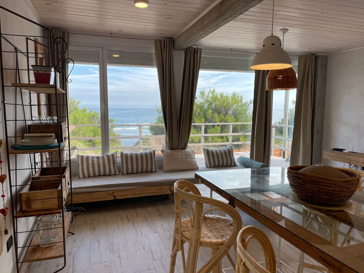 A bright living space features large windows that reveal a scenic ocean view. A comfortable bench with striped cushions lines the wall, and a wooden dining table with stylish chairs occupies the center. Natural light filters in through sheer curtains.