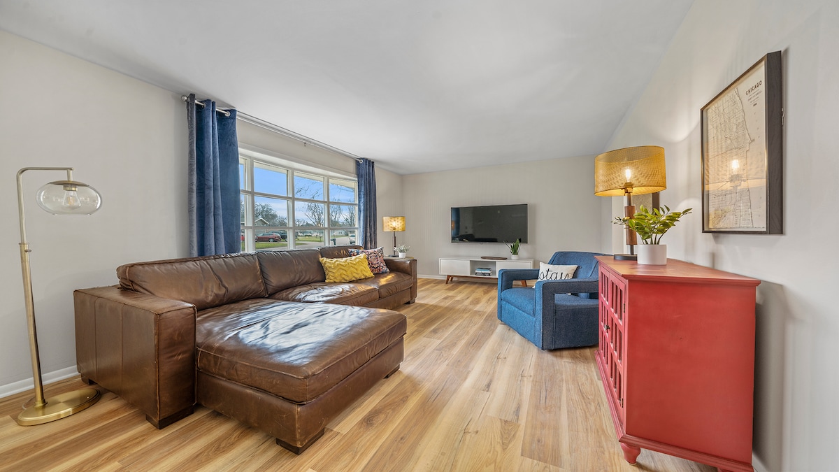 A cozy living area features a brown leather sectional sofa paired with a blue accent chair. Two lamps with soft lighting sit atop side tables. A television is mounted on the wall across from the seating, and natural light enters through large windows, highlighting the wooden flooring.