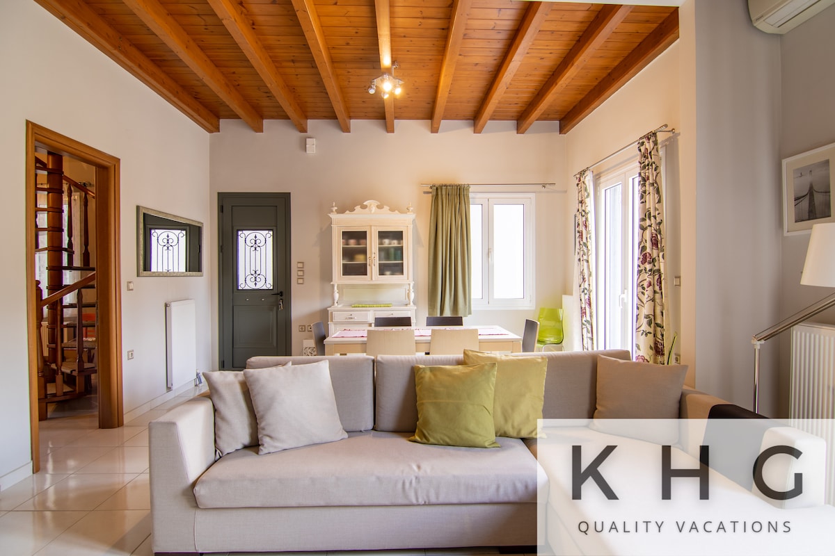 A spacious living room features a comfortable sofa with soft cushions, complemented by wooden beams on the ceiling. A dining area is visible, illuminated by natural light from large windows adorned with patterned curtains. An internal staircase can be seen in the background.