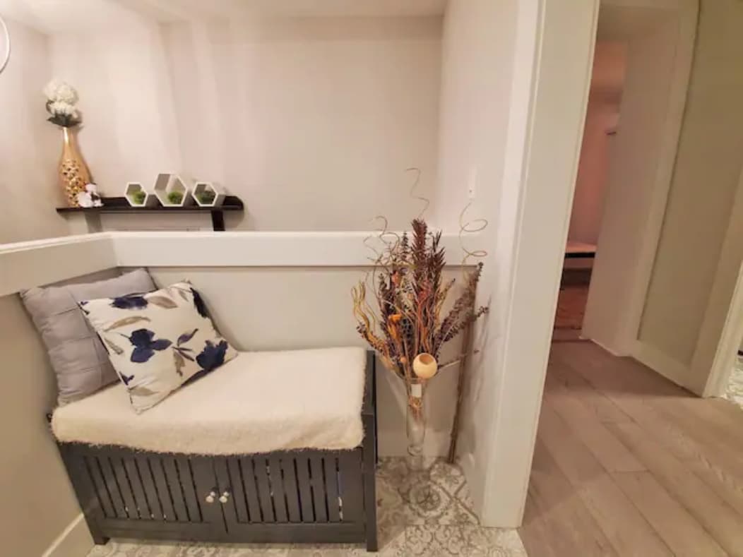 A cozy entryway nook features a cushioned bench adorned with a floral pillow, while a decorative arrangement of dried branches and a small vase add a touch of elegance. The light and neutral tones of the walls create a welcoming atmosphere.