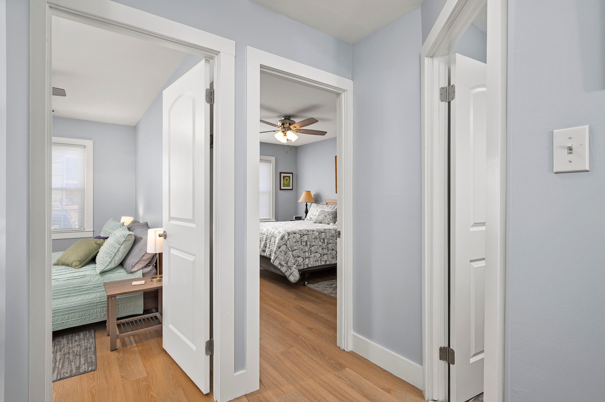 A hallway connects two bedrooms, featuring light blue walls and hardwood flooring. One room contains a spacious bed with a soft green blanket, while the other showcases a queen bed with a floral comforter. Natural light filters through the nearby windows.