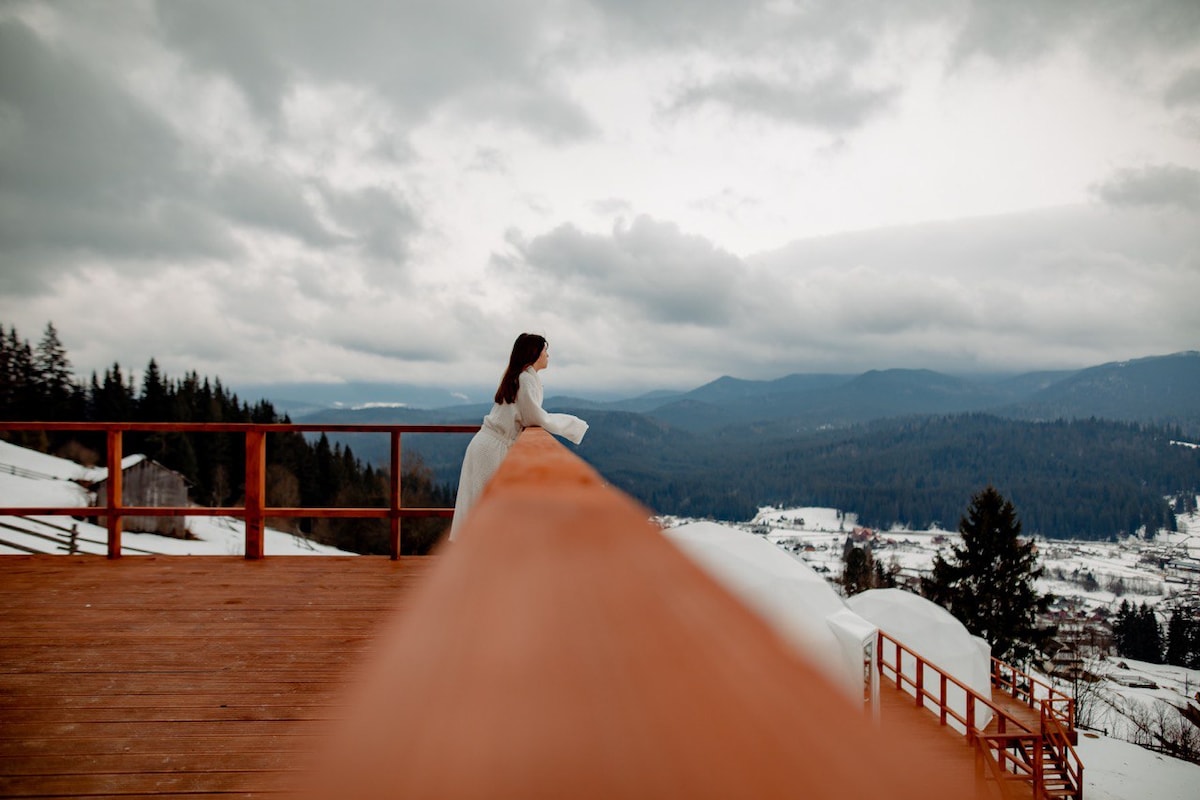 A wooden railing extends along a balcony, providing an unobstructed view of the surrounding mountains and valleys. Cloudy skies create a dramatic backdrop, while a person is seen gazing towards the horizon, dressed in a light-colored outfit that gently contrasts with the snowy landscape.