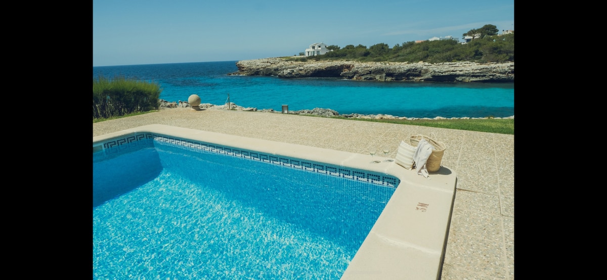 A sparkling pool showcases clear blue water, extending toward the coastline. The surrounding area features well-maintained grass and rocky shorelines, with the ocean’s vibrant hues of blue visible in the background under a clear sky.