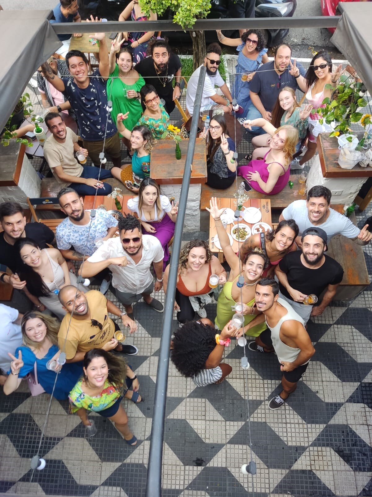 A vibrant outdoor gathering is depicted from above, showcasing a lively community atmosphere. Tables are filled with food and drinks, surrounded by guests enjoying their time together. Decorative greenery adds a touch of nature to the festive scene.