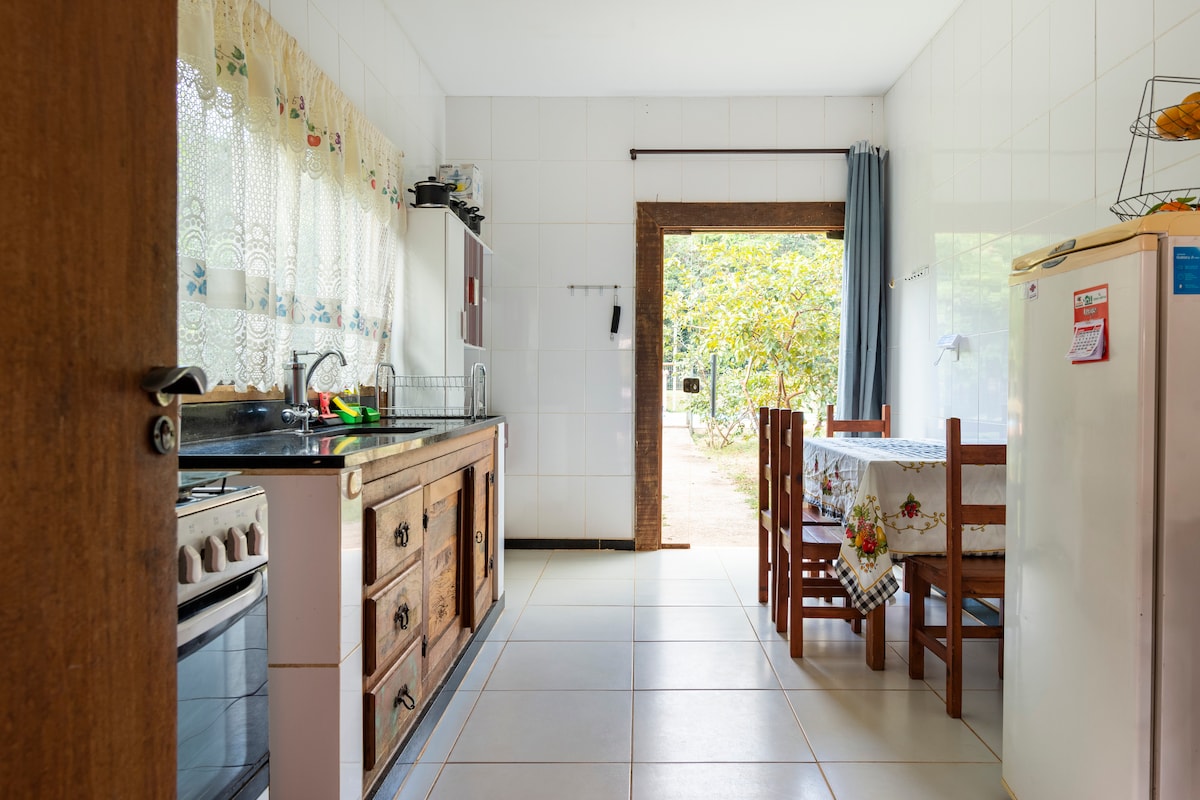 A kitchen and dining area feature wooden cabinets and a simple dining table with four chairs. Natural light flows through a door leading to the outdoor space. Counters are equipped with essential appliances, including a stove and refrigerator.