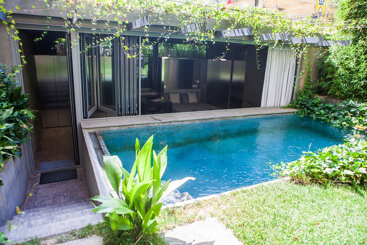 A serene outdoor pool is framed by lush greenery and stone accents. The contemporary architecture of the adjoining space features large glass doors that open to reveal a spacious interior. Nature is intertwined with the design, creating a tranquil retreat.