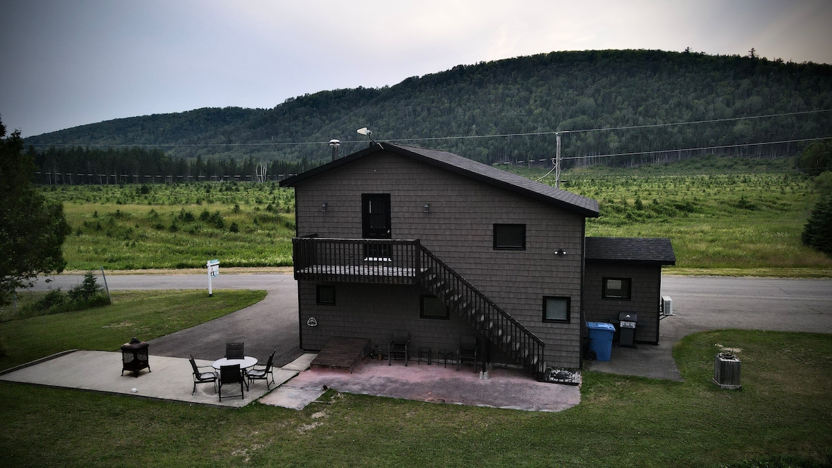The two-story cottage features a dark exterior and a balcony overlooking the surrounding landscape. A patio area with several outdoor chairs is seen, while a grassy field extends towards a wooded hill in the background.