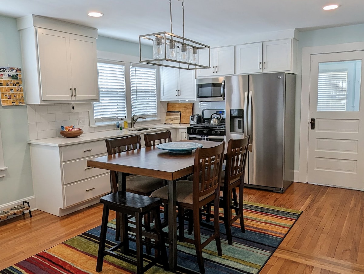 The kitchen features updated appliances, including a stainless steel refrigerator and stove. A wooden dining table with six chairs sits on a colorful striped rug, enhancing the open floor plan. Natural light enters through the windows, illuminating the bright, airy space.