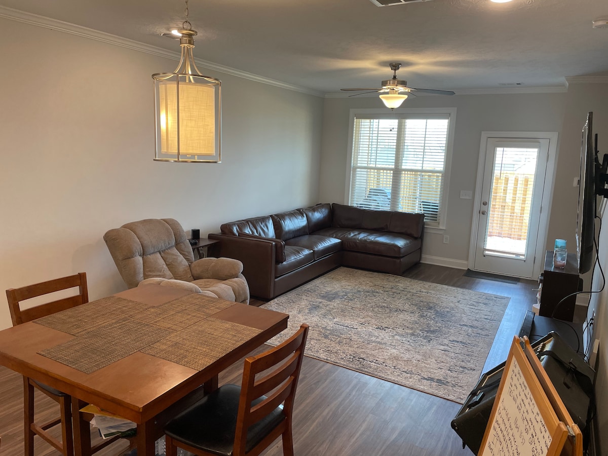 A spacious living area features a comfortable brown sectional sofa, a recliner, and a wooden dining table with four chairs. Natural light enters through the windows, providing a bright ambiance. A door leads to a private outdoor space, complementing the open layout.