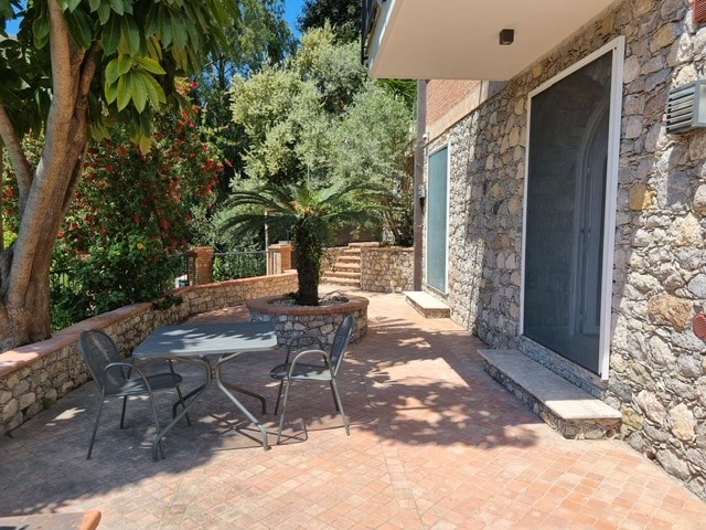 A sunlit outdoor area is framed by lush greenery and colorful flowers. A stone pathway leads to a circular seating area, furnished with a table and several chairs, inviting relaxation in a serene garden setting.
