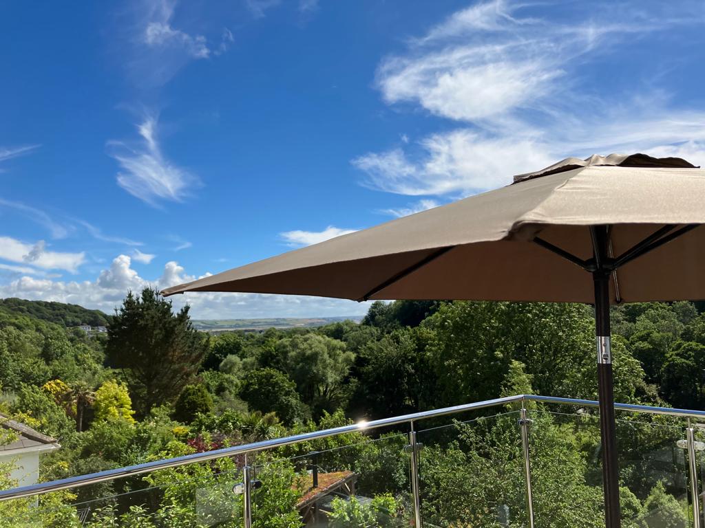 A spacious rooftop balcony is shown, featuring a large umbrella providing shade. Lush greenery and trees are visible in the landscape, with clear blue skies overhead. The glass balustrade offers an unobstructed view of the valley beyond.
