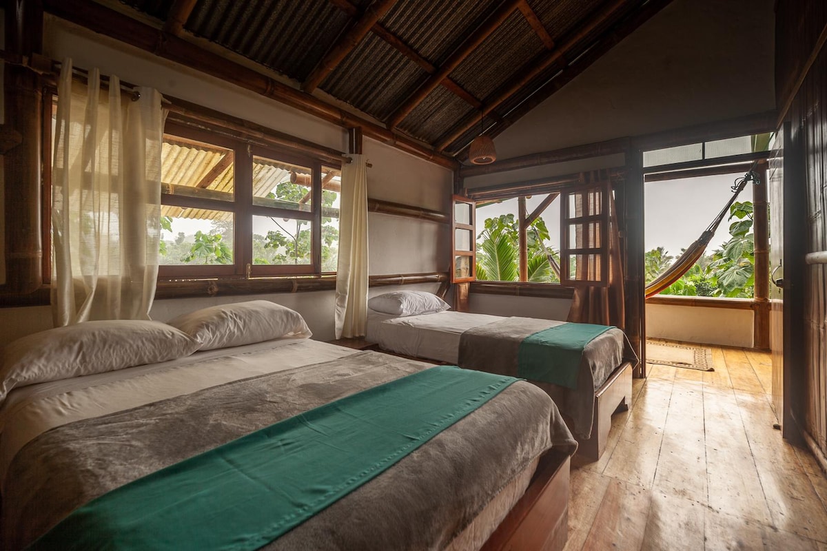 A spacious room is furnished with three beds, each dressed in soft linens. Large windows with sheer curtains allow natural light to fill the space and provide views of lush greenery outside. A hammock is seen in the corner, enhancing relaxation options.