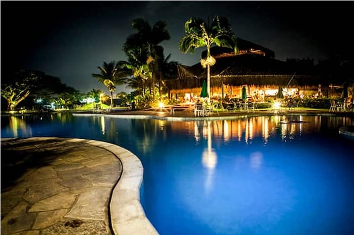 The illuminated pool reflects the soft glow of surrounding lights, accompanied by palm trees gently swaying in the breeze. The area suggests a serene atmosphere for relaxation, with seating available nearby for guests to enjoy the tranquil setting.