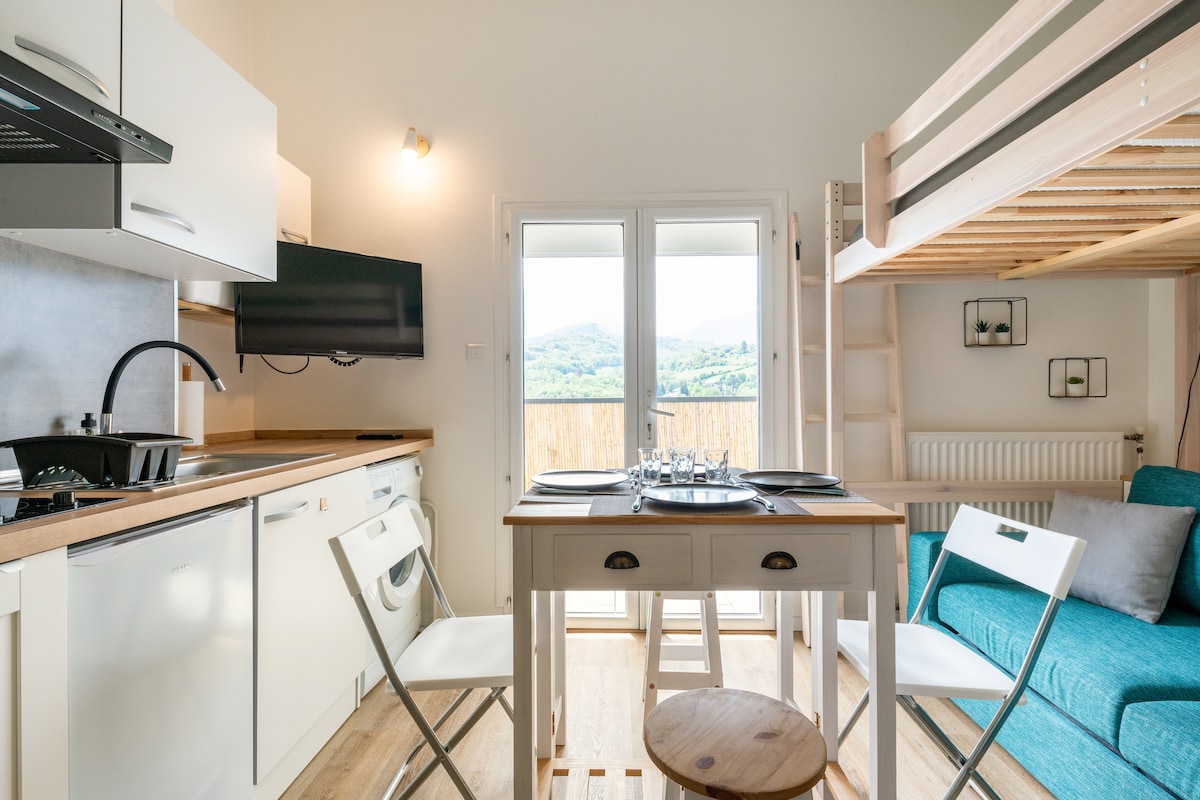 A modern kitchen area is equipped with essential appliances, including a stovetop and a television mounted on the wall. A dining table is set for four, surrounded by white chairs. Natural light enters through double doors, offering scenic views of the landscape beyond.