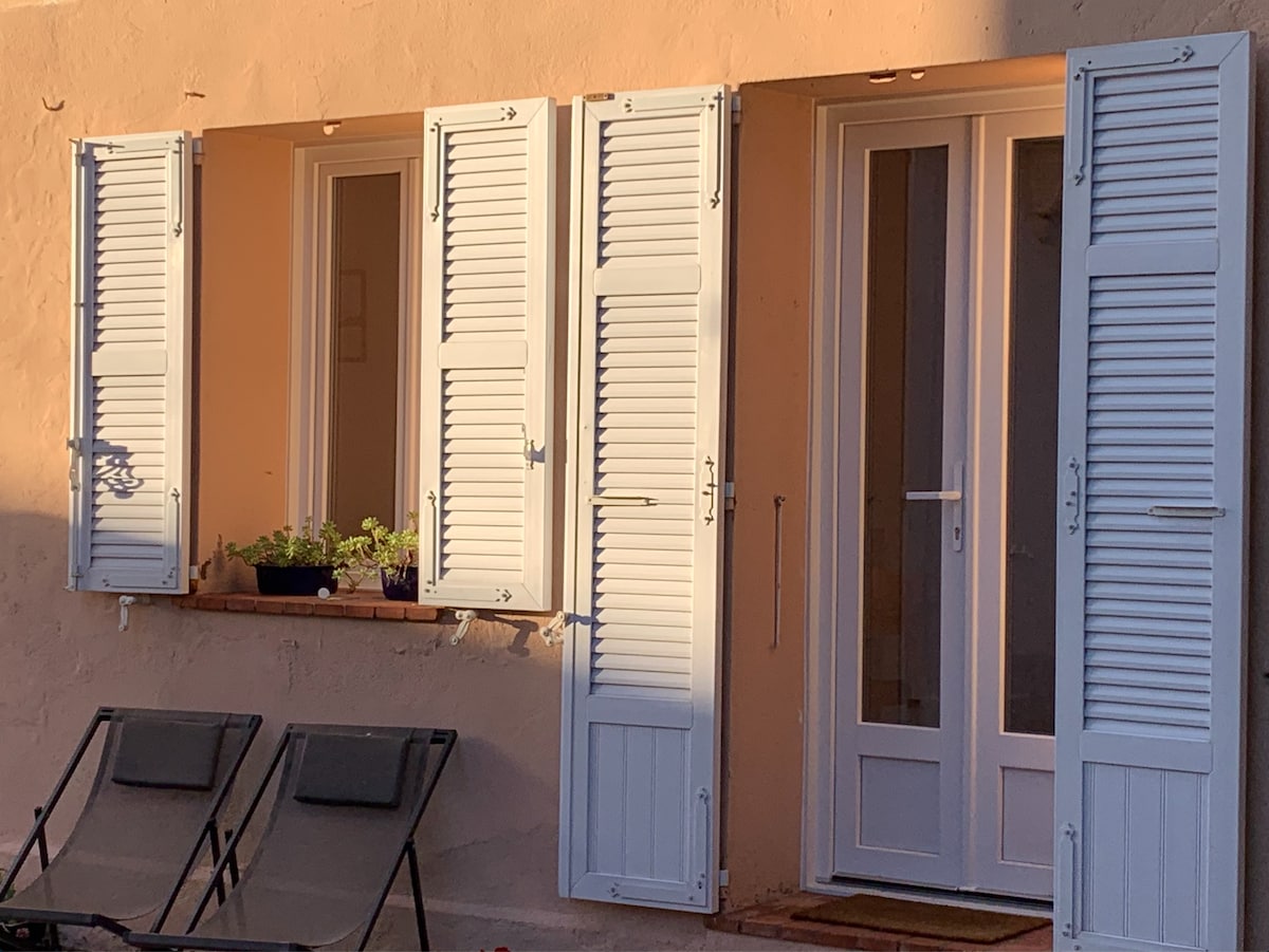 A charming exterior view displays two sets of white shutters flanking a glass door and window, which open to the outside. A low ledge holds potted plants, while two reclining chairs are positioned nearby, inviting relaxation in a serene environment.