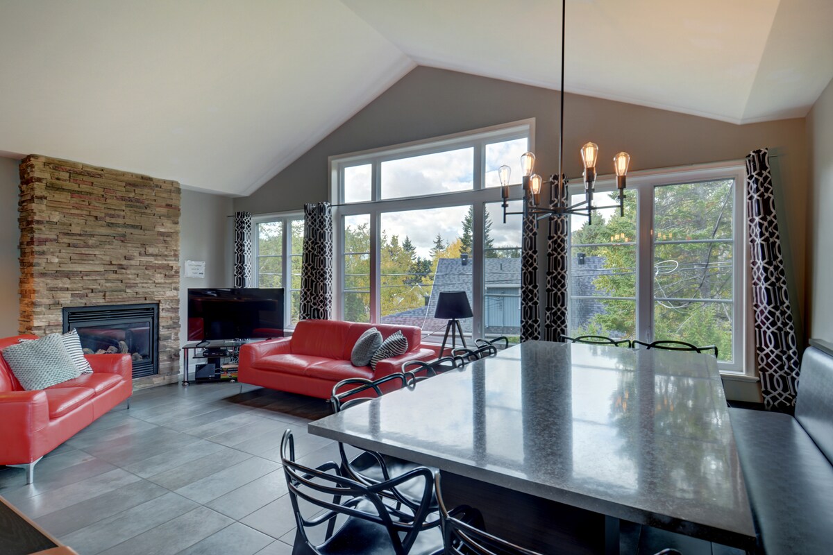 The living and dining area features high ceilings and large windows, allowing natural light to fill the space. A modern, rectangular dining table is surrounded by black chairs. Red leather sofas provide comfortable seating next to a stone fireplace, creating a welcoming gathering area.