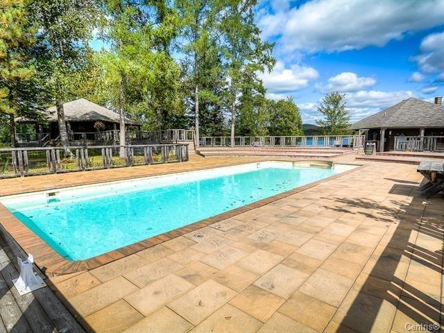 A spacious outdoor pool is surrounded by a wooden deck and shaded by trees. Sunlight reflects off the calm water, and nearby structures can be seen in the background. The setting provides a relaxed atmosphere for leisure activities.