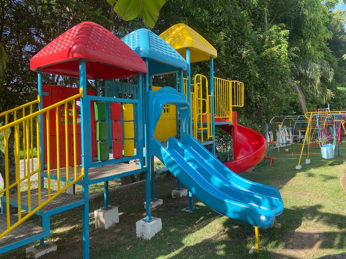 A colorful playground setup features a bright blue slide, yellow and red play structures, and multicolored panel walls. Lush greenery surrounds the area, creating a vibrant outdoor space suitable for children. Additional play equipment can be seen in the background.