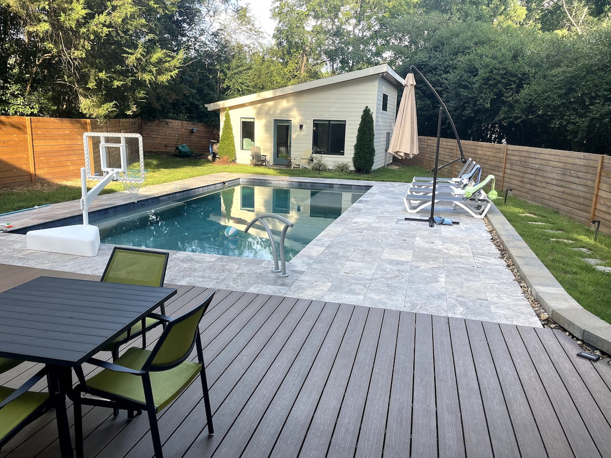 A swimming pool is centrally located in the backyard, surrounded by a tiled deck. Lounge chairs line one side, while a patio table with green chairs is positioned nearby. The guesthouse is seen in the background, flanked by trees and a wooden fence.
