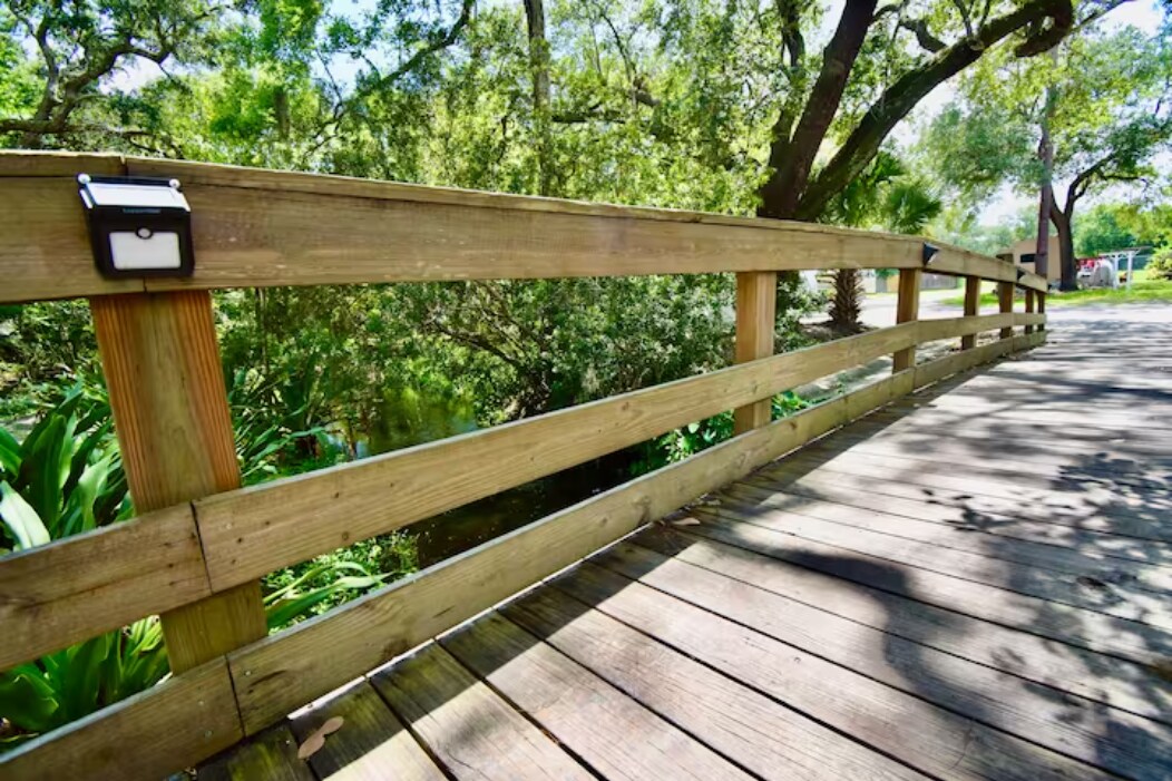 A wooden bridge extends across a lush, green area, lined with trees and shrubs. Sunlight filters through the foliage, creating gentle patterns on the wooden planks. The sturdy railing, equipped with subtle lighting, enhances visibility along the bridge.