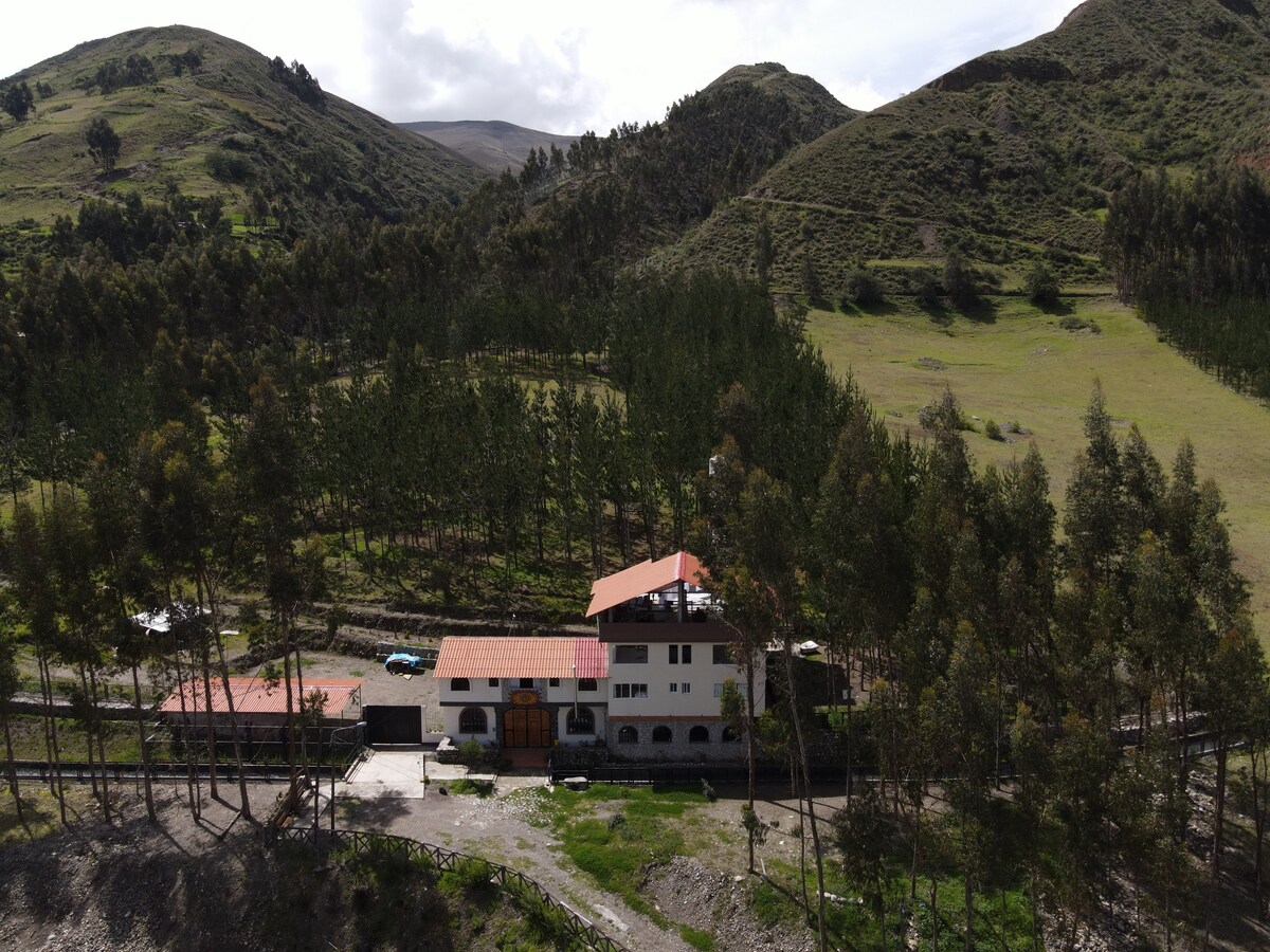 The ecolodge is nestled among rolling green hills, surrounded by tall trees. Its architecture displays a multi-level structure with a reddish-brown roof and large windows. A parking area is visible beside the building, enhancing accessibility to the scenic environment.