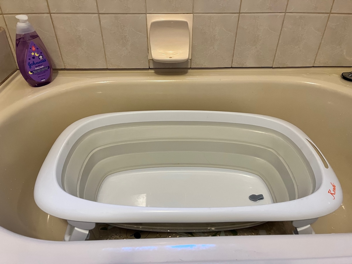 A white, foldable bathtub is positioned inside a regular bathtub, featuring a simple design with a smooth surface and a small footed base. A purple bottle of liquid soap is visible on the side, adding functionality to the bathing space.