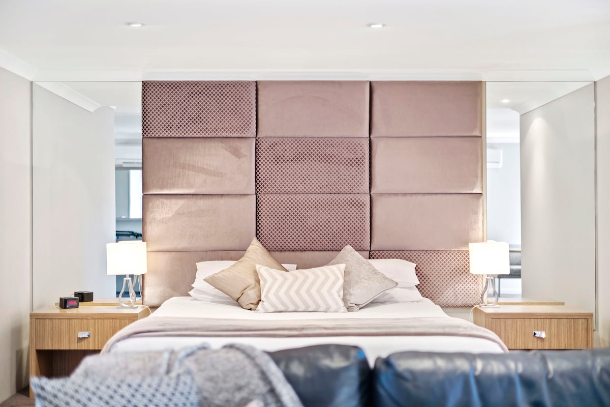 A spacious sleeping area features a stylish padded headboard in soft, muted tones. On either side of the bed, modern bedside tables are illuminated by warmly glowing lamps. Coordinated cushions rest on the bed, enhancing the serene ambiance of the room.