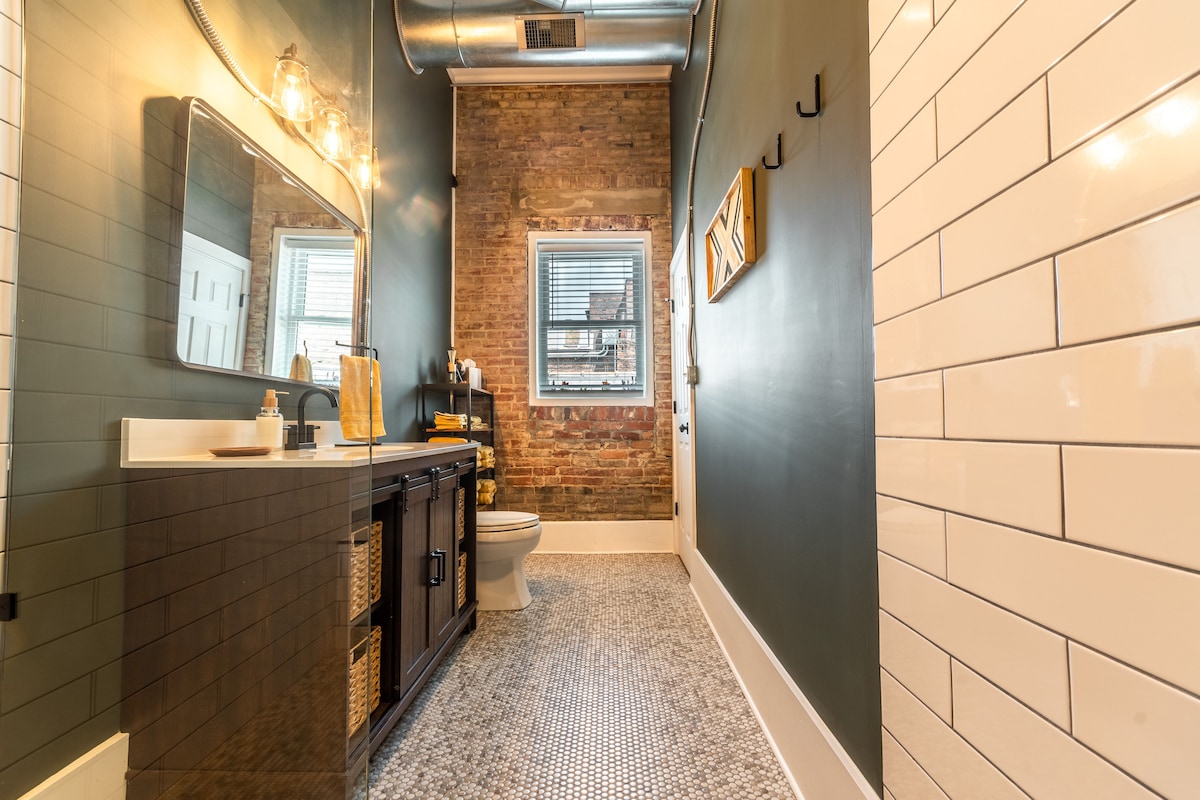 A well-lit bathroom features a combination of exposed brick and smooth white tiles. A large mirror hangs above a dark vanity with storage baskets underneath. Natural light is provided by a window with light-filtering blinds, while the flooring showcases subtle patterned tiles.