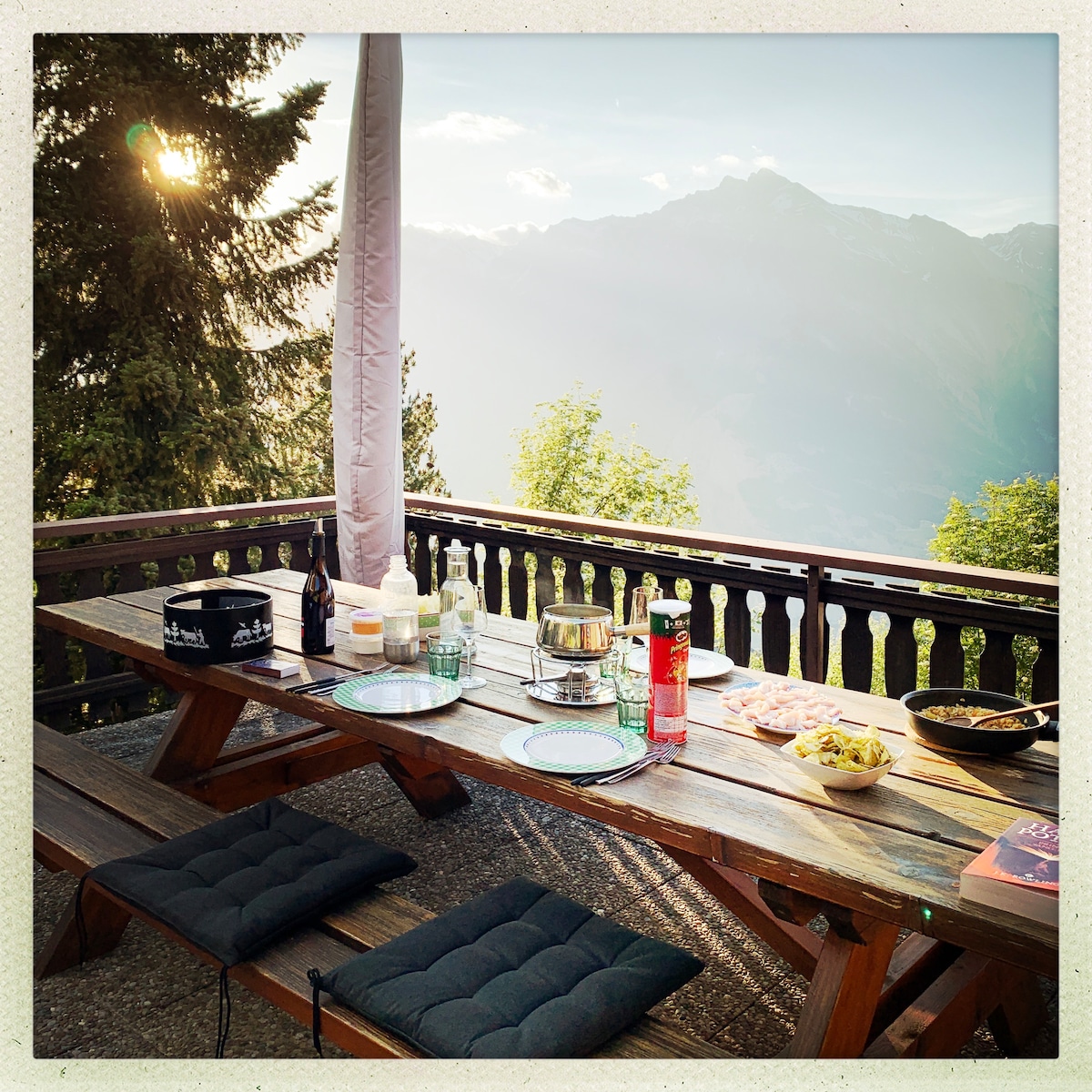An outdoor dining area features a large wooden table surrounded by benches. Various beverages and food items are arranged on the table, with a scenic mountain view in the background, characterized by soft sunlight filtering through the trees.