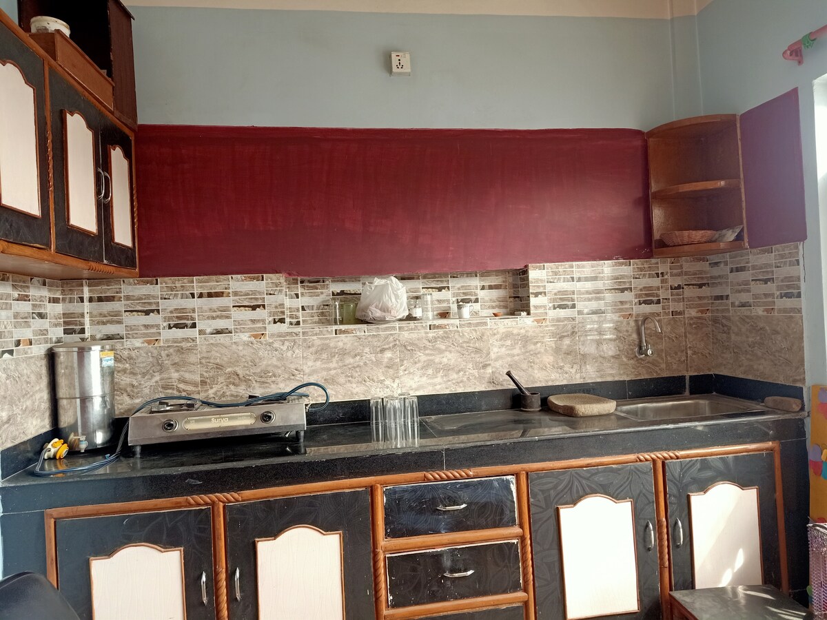 A functional kitchen features dark wooden cabinetry with cream-colored accents. A stone countertop is complemented by a tiled backsplash. Cooking appliances, including a stovetop and utensils, are visible, while natural light filters in through a window, illuminating the space.
