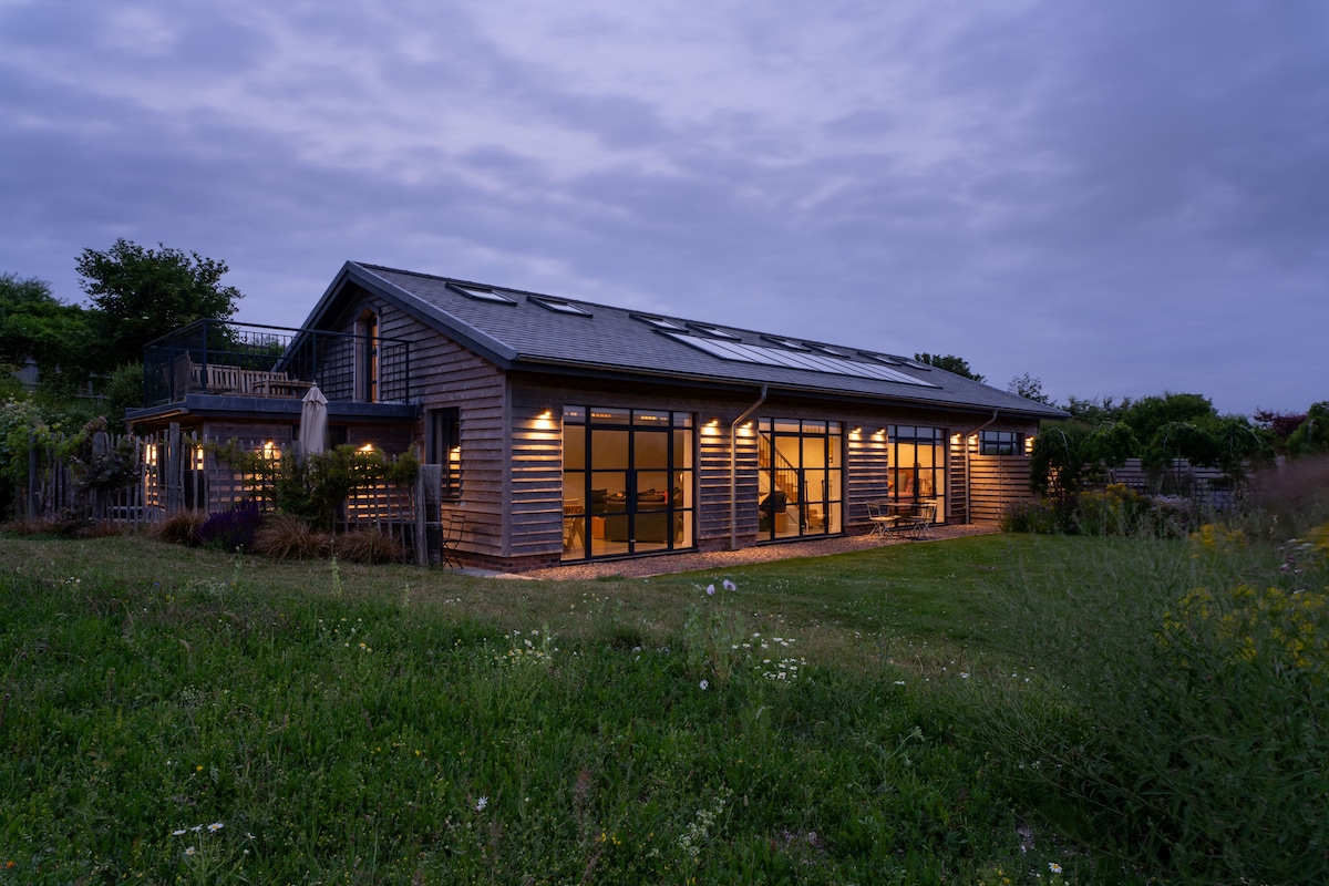 The exterior of the eco-converted Grain Store is showcased during twilight. The building features a modern design with large windows, illuminating the interior space, while the surrounding lush greenery and low shrubbery enhance the serene ambiance.