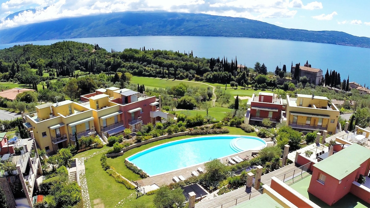 An aerial view showcases a scenic residential complex with colorful buildings surrounding a serene, curved swimming pool. Lush gardens and well-kept greenery enhance the tranquil atmosphere. In the background, the expansive Lake Garda glistens under the sky, framed by rolling hills and distant mountains.