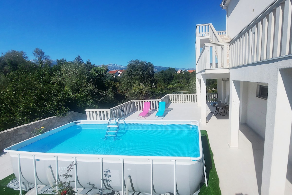 A rectangular above-ground pool occupies the center of a sunlit patio, surrounded by green grass. Colorful lounge chairs in shades of pink and blue are positioned nearby, while a small table and chairs are situated on a covered terrace. Lush greenery is visible in the background.