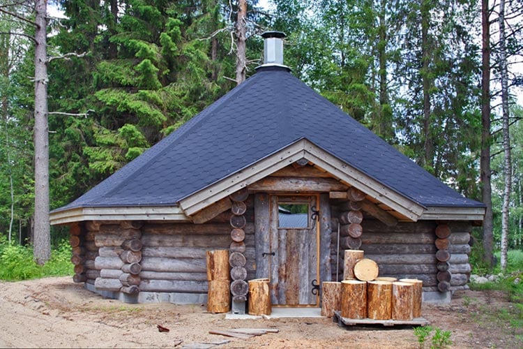 A rustic log cabin features a distinctive conical roof and a sturdy wooden door. Logs are neatly arranged outside, providing an accessible storage option. The surrounding forest provides a natural backdrop, enhancing the serene setting for gatherings and outdoor activities.