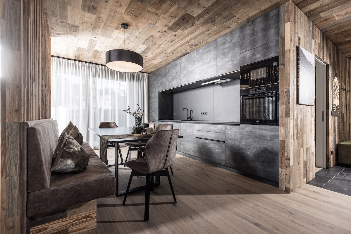 An open-plan kitchen and dining area showcases elegant wood paneling and modern cabinetry. Soft fabric seating surrounds a rectangular dining table. Large windows allow natural light to flood the space, with a pendant light offering additional illumination overhead.