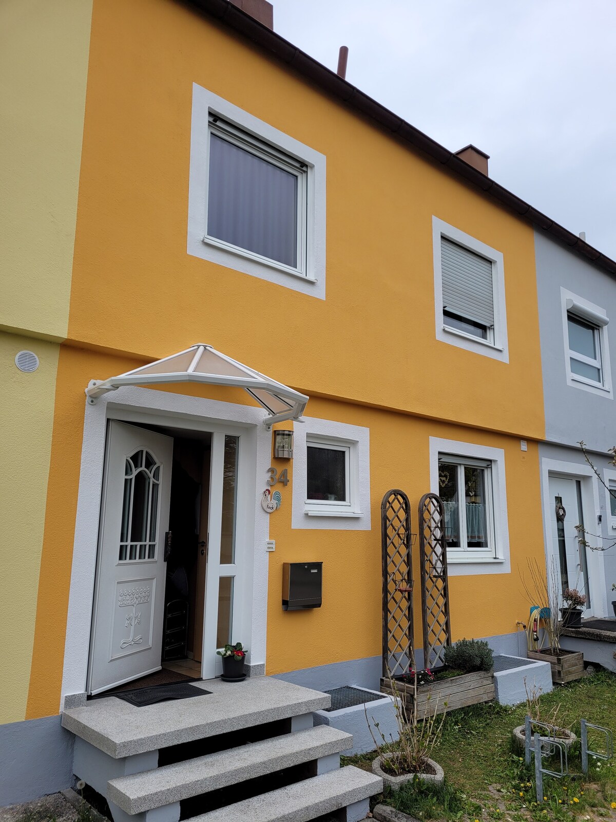 The exterior of a two-story townhouse is shown, featuring a bright yellow facade with white trim. A covered entrance leads to the door, flanked by decorative trellises. Steps provide access, while a mailbox is mounted beside the entryway.
