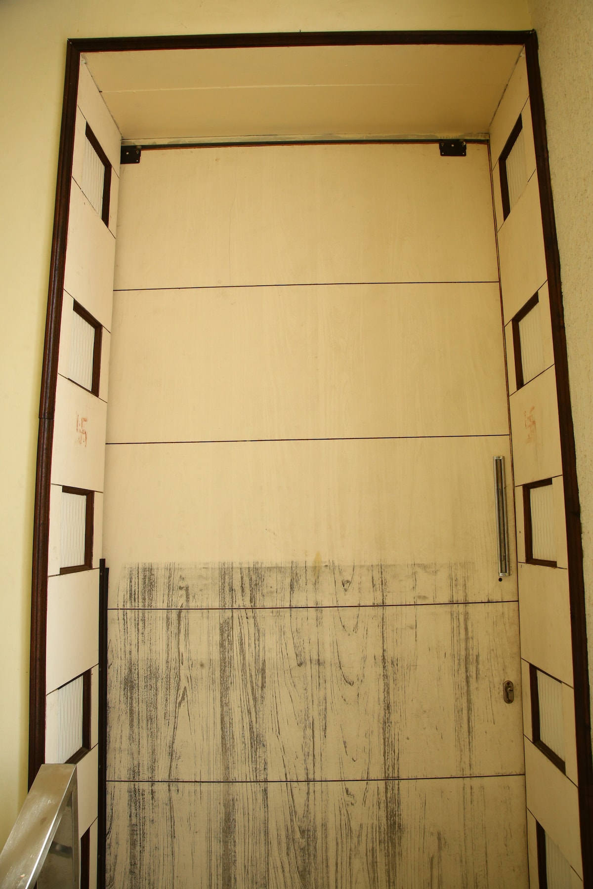 A plain wooden door is framed by beige walls, featuring a simple design with vertical panels. A metal handle is positioned on the right side. Soft light is visible, indicating an indoor space beyond the door.
