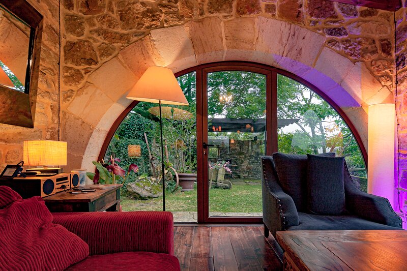 A spacious living area is highlighted by a large arched window that offers views of a well-maintained garden. Soft lighting from table lamps enhances the inviting atmosphere. Comfortable seating includes a plush couch with a textured design and an armchair, creating a cozy space for relaxation.