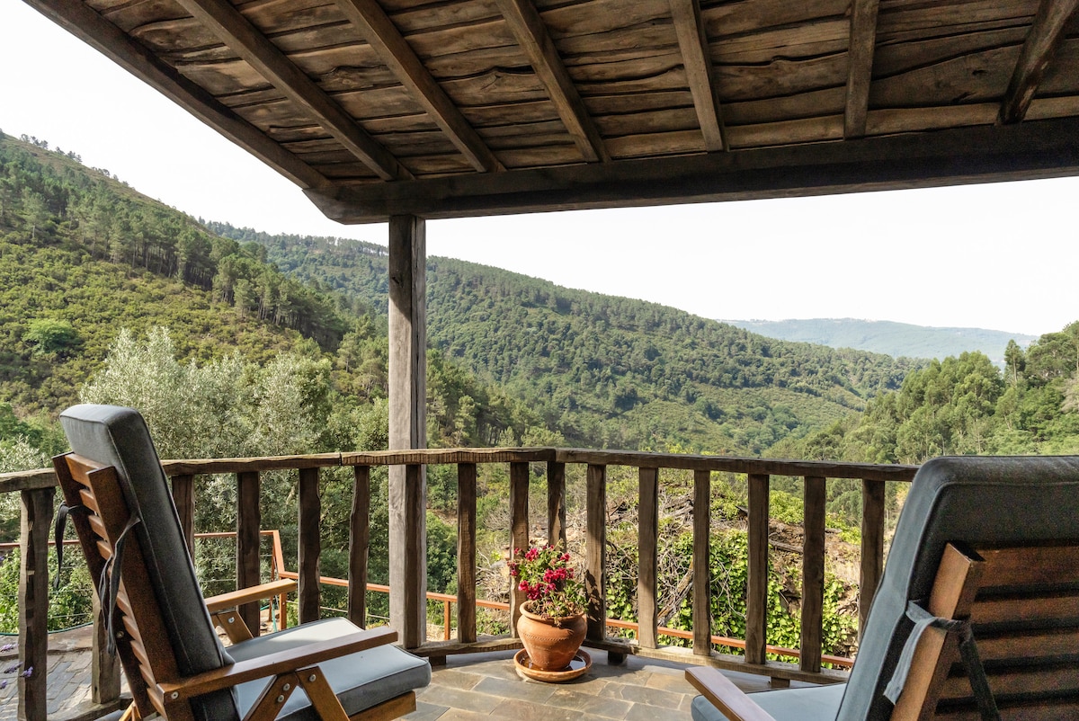 A spacious outdoor terrace features wooden railings and two lounge chairs, offering a serene view of the lush valley and surrounding hills. A potted plant with vibrant flowers adds a touch of color, complementing the natural landscape.