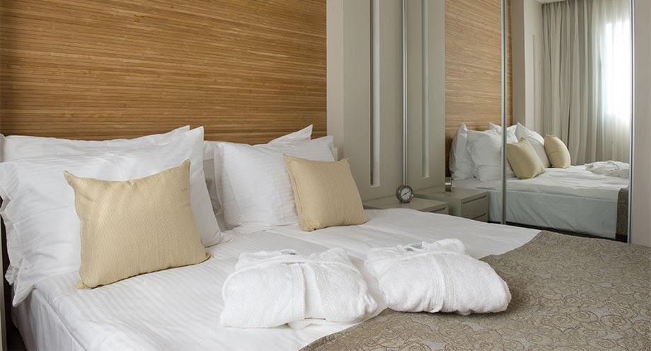 A bright sleeping area is presented, featuring a spacious bed adorned with white linens and two decorative pillows. Soft towels are neatly placed at the foot of the bed. A mirrored closet reflects the light from the window, enhancing the room's openness.