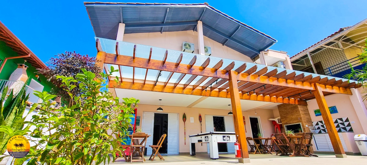 An outdoor view of a two-story building is presented, featuring a wooden pergola outfitted with seating. Air conditioning units are mounted on the upper floor, and vibrant plants frame the exterior, enhancing the inviting atmosphere of the property.