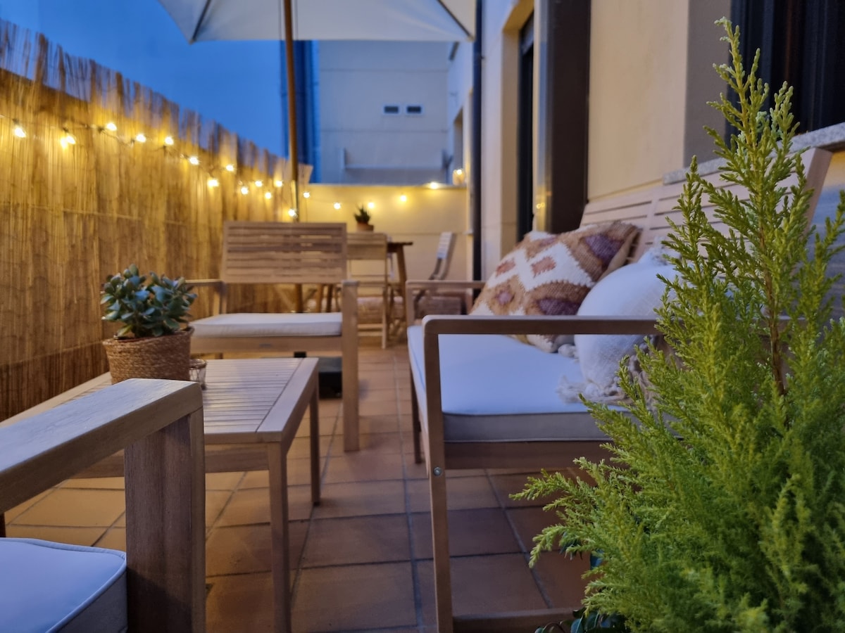 An outdoor terrace is equipped with wooden furniture, including seating and a small table, surrounded by soft lighting. Potted plants add greenery to the space, creating a cozy atmosphere for relaxation. An umbrella offers shade, enhancing the usability of the terrace.